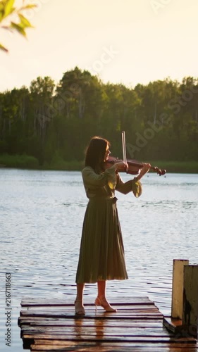 Woman playing violin on rustic wooden pier at tranquil lakeside during golden hour sunset, surrounded by calm water and lush forest landscape