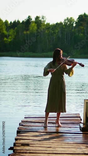 Young woman playing violin on rustic wooden pier over tranquil lake, surrounded by lush green forest and soft natural light at peaceful golden hour