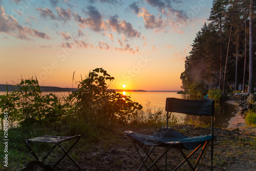 Beautiful sunset over a peaceful lake surrounded by forest, with camping chairs and a campsite overlooking the water. High quality photo