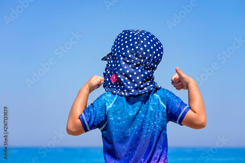 Close up of a girl looking at the sea, Child wearing rash vest and a hat to protect form the sun, skin cancer awareness and protection. girl wearing Sunsafe Swimsuit providing UVA +UVB protection