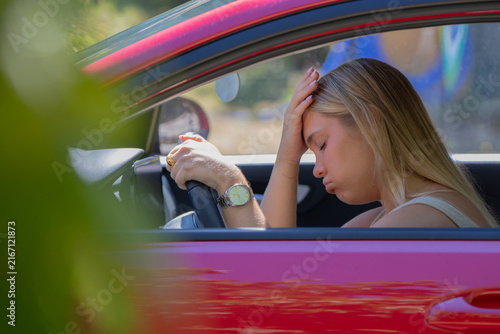 Young woman sitting in car with hand on forehead, looking stressed and exhausted while holding steering wheel