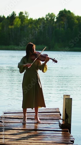 Young woman playing violin on a rustic wooden pier by a calm lake, surrounded by lush green forest and soft afternoon light in a peaceful outdoor setting