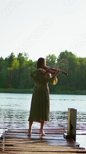 Woman in flowing green dress playing violin on rustic wooden pier by tranquil forest lake, captured in peaceful outdoor sunset performance sequence