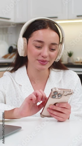Young woman at home wears wireless headphones types message on smartphone smiles takes break from work. Caucasian girl on sofa sits at table with laptop checks social media chats relaxes calmer mood