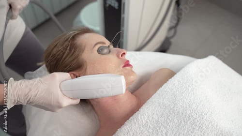 A woman lies on a treatment bed receiving a facial laser procedure. She wears protective eye shields while a technician applies a handheld device to her jawline