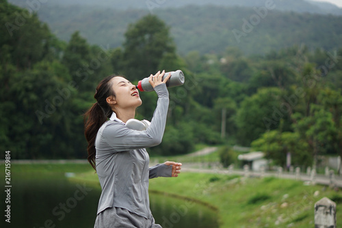 A side profile shot of a young Asian woman drinking water from a sports bottle to stay hydrated during her outdoor fitness session by the lake.