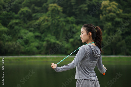 Side view of a sweaty young Asian woman standing by a lake holding a skipping rope behind her back after an intense workout session.