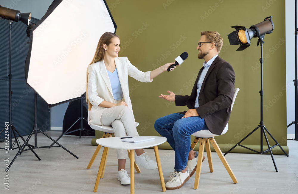 Obraz premium Interview with journalist and guest in studio. A woman interviewer holds a microphone to a man as lights and softbox shape pro media set for talk. Clear media reporter interview concept.