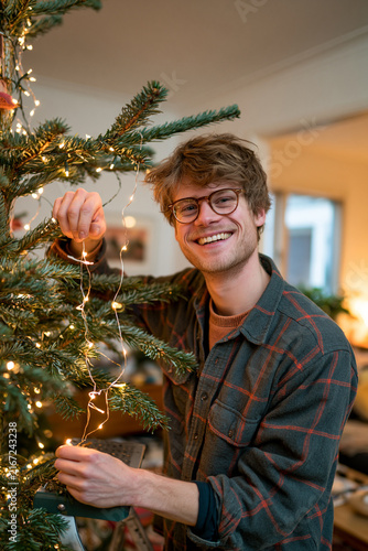 Happy young man in glasses decorating Christmas tree with glowing garland lights at home during holiday season