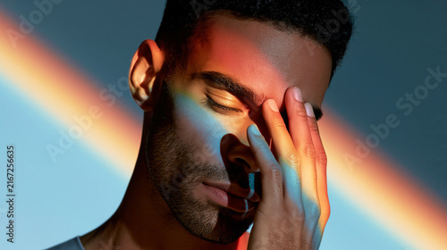 Young man with eyes closed touching forehead with rainbow light symbolizing identity and self acceptance.