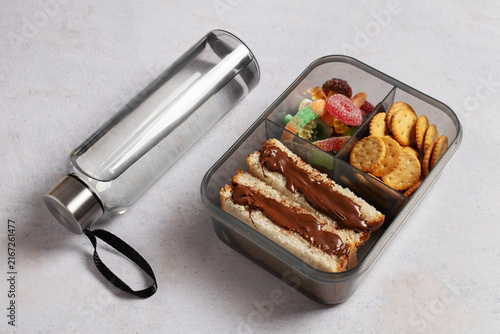Unhealthy lunchbox with chocolate spread sandwich, crackers and gummy candy next to water bottle. Sugar and ultra-processed food kids nutrition concept.