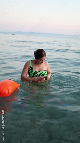 Vertical screen woman attaches bright orange swim safety buoy to waist in shallow sea preparing for safer visible swimming during summer vacation along the Croatian coast