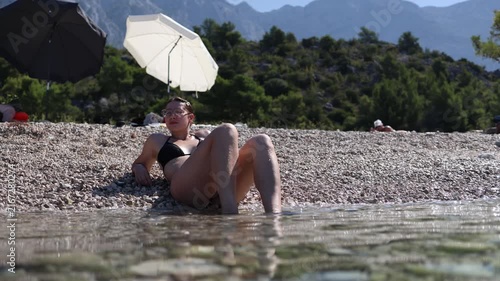 Woman relaxes at the waters edge on a pebble beach wearing protective water shoes while enjoying a sunny summer vacation with dramatic mountain scenery rising behind hers