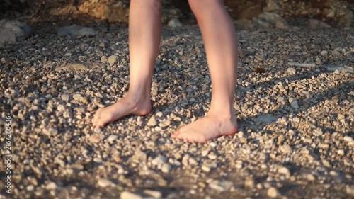 Close view of a woman walking barefoot over uneven pebbles on a rocky beach lifting each foot carefully as rough stones make every step uncomfortable by the sea at sunset