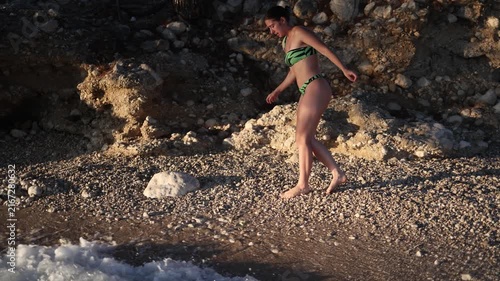 Woman struggles to walk barefoot across sharp stones on a rocky beach because she has no protective shoes while carefully approaching the ocean during her summer vacation