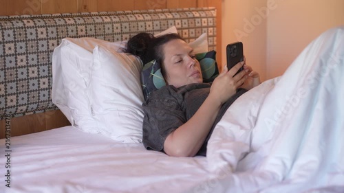 Woman is lying in bed covered with a blanket, texting on the phone. Girl under the blanket sends a message with his smart phone and smiles because he received an amusing message or love, and responds 