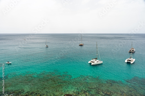 Aerial view of multiple sailboats and catamarans anchored in turquoise ocean water.
