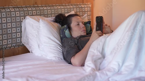 Woman is lying in bed covered with a blanket, texting on the phone. Girl under the blanket sends a message with his smart phone and smiles because he received an amusing message or love, and responds 
