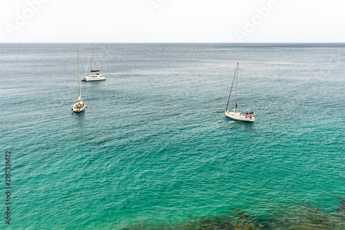 Aerial view of sailboats and catamaran anchored in turquoise sea with swimmers nearby.