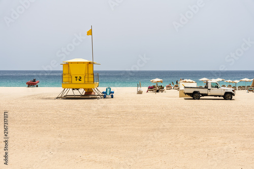 Yellow lifeguard tower with flag on sandy beach beside truck and turquoise sea in Morro Jable, Fuerteventura.