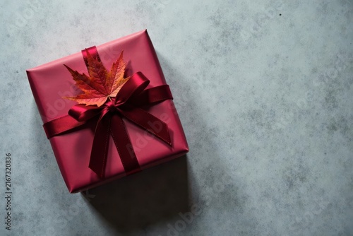 A Crimson Gift Box Adorned with an Autumn Leaf and a Rich Burgundy Ribbon, Resting on a Textured Surface