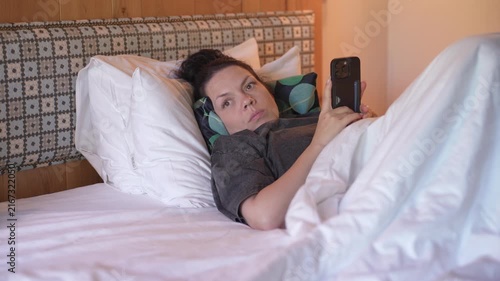 Woman is lying in bed covered with a blanket, texting on the phone. Girl under the blanket sends a message with his smart phone and smiles because he received an amusing message or love, and responds 