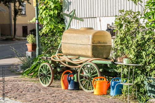 Historischer Leiterwagen aus Holz mit Speichenrädern und Wassertank aus Kunststoff und farbigen Gießkannen vor ländlichem Gebäude mit Holzwand und Kopfsteinpflaster in Bempflingen.