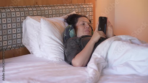 Woman is lying in bed covered with a blanket, texting on the phone. Girl under the blanket sends a message with his smart phone and smiles because he received an amusing message or love, and responds 