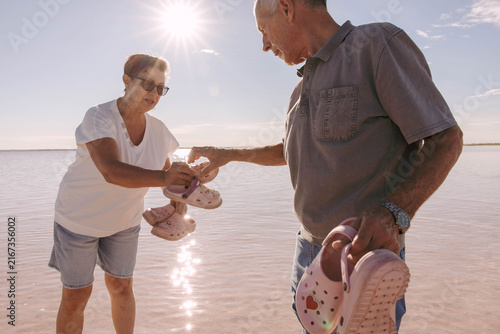 Senior grandparents carry their grandchildren's shoes while walking through the shallow waters of a pink salt lake on a sunny summer day. care, support, active aging, multigenerational travel