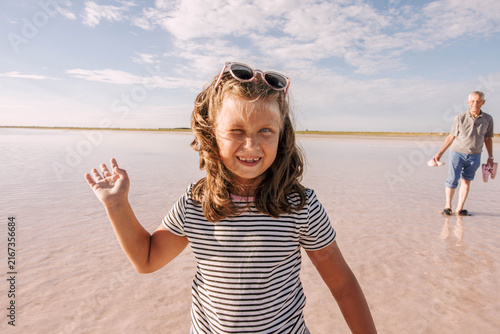 little girl smiling waving in shallow pink salt lake. Grandfather carrying children's shoes. Sunny summer day, playful family moment, travel, childhood and outdoor lifestyle.