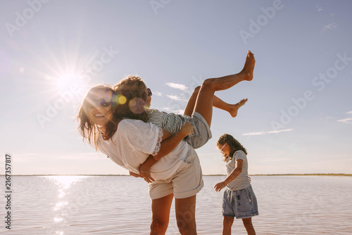 smiling mother carrying playful daughter at shallow pink salt lake, sunny family vacation with another child walking nearby, joyful outdoor travel scene, family tourism, family resort, parenting