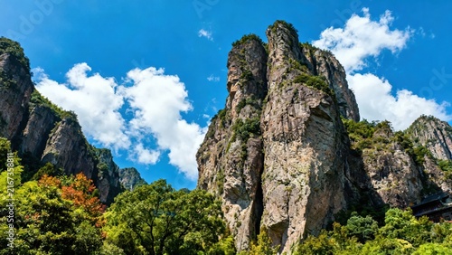 Towering rock formations rise against a clear blue sky, surrounded by lush green trees and vegetation in a mountainous landscape.