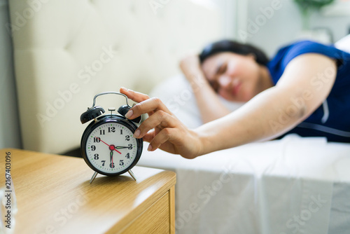 Cuadro en lienzo Sleepy woman waking up to a ringing alarm clock, extending hand to snooze or tur