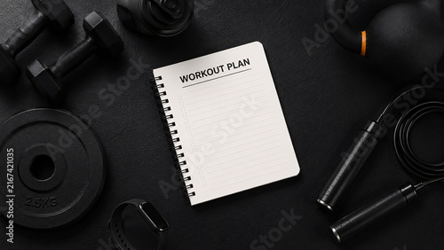 Flat lay top view of blank workout plan notebook surrounded by dumbbells kettlebell jump rope and fitness accessories on dark background