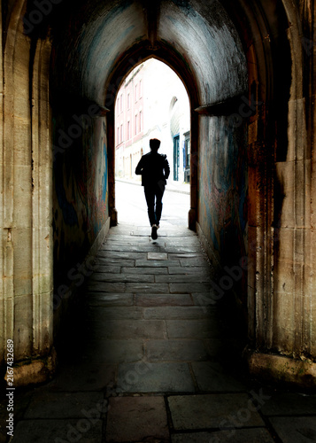 A silhouette of a person running through a dark, arched stone tunnel towards a bright city street, capturing a sense of transition and forward momentum.