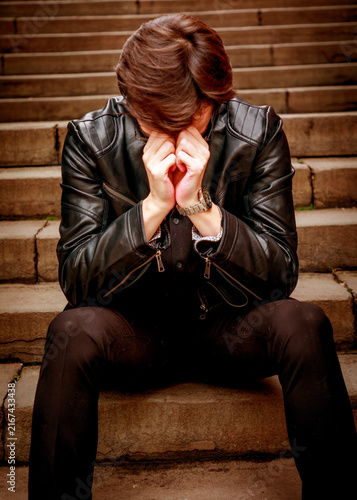 Foto A young man in a black leather jacket sits alone on outdoor stone steps, head bowed and hands covering his face, capturing a poignant moment of deep reflection or emotional exhaustion