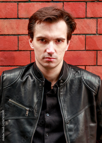 A portrait of a young man with brown hair, dressed in a black shirt and leather jacket, posing confidently against a bold red brick wall background.