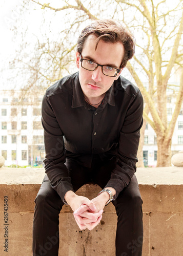 A man in a black shirt and glasses sits calmly on a stone ledge outdoors, looking directly into the camera during a portrait photography session.