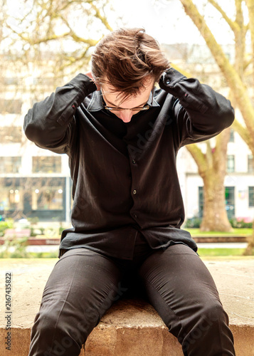 A young man in a black shirt sits outdoors with his hands behind his head and his gaze cast downward, seemingly lost in quiet contemplation or taking a moment to rest in the park.