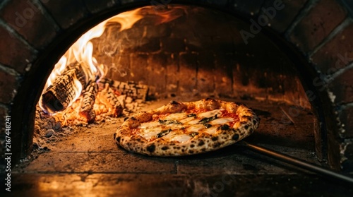 Fresh neapolitan pizza baking inside traditional brick wood fired oven with bright orange flames and glowing embers background.