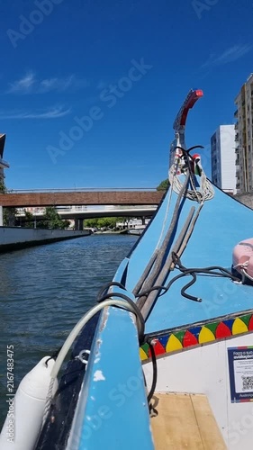 POV shot from the prow of a traditional colorful Moliceiro boat gliding along the calm water of a canal in Aveiro, heading towards a bridge under a clear blue sky in Portugal.