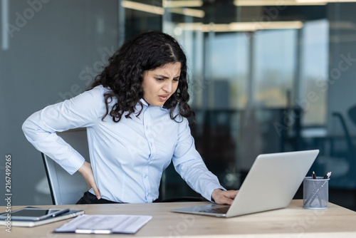 Foto Young woman feeling severe low back pain while sitting at her office desk and wo
