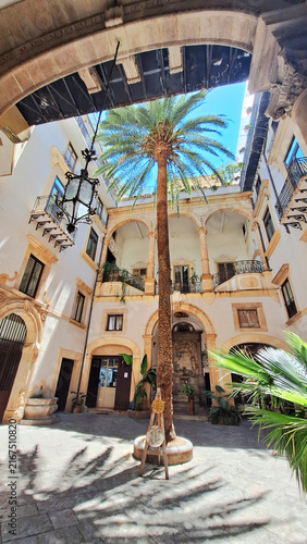 Vertical shot of the beautiful baroque courtyard inside Palazzo Castrone in Palermo, Sicily, Italy, featuring a tall centennial palm tree, elegant stone loggias, and an old wrought iron lantern.