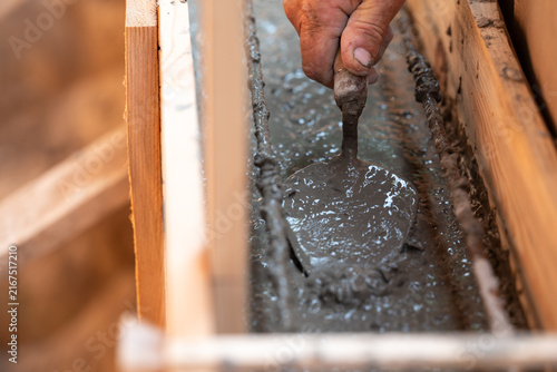 Builder smoothing concrete with a trowel in wooden formwork. A worker's hand dips a trowel into fresh concrete mortar inside formwork, symbolizing the foundation construction process