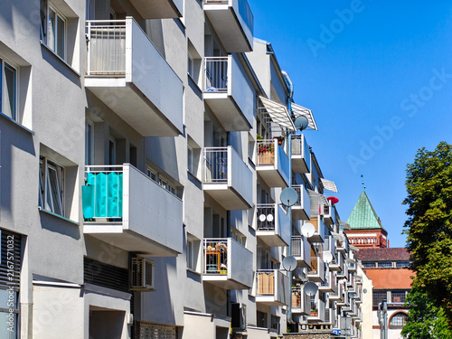 Фотография Modern residential building facade with numerous balconies and satellite dishes against a clear blue sky, with a view of a distant red brick church with a green copper roof and a mature tree