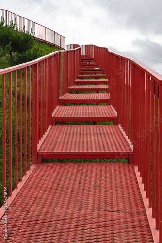 Картината върху платно Red metal staircase and walkway climbing a green hillside under cloudy sky