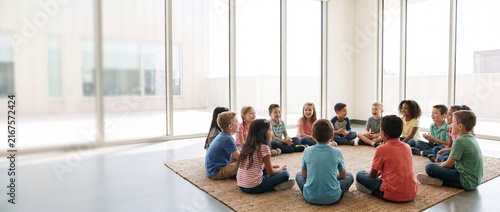A diverse group of young children sitting in a circle on a large rug in a bright, modern kindergarten or daycare playroom with large floor-to-ceiling windows.