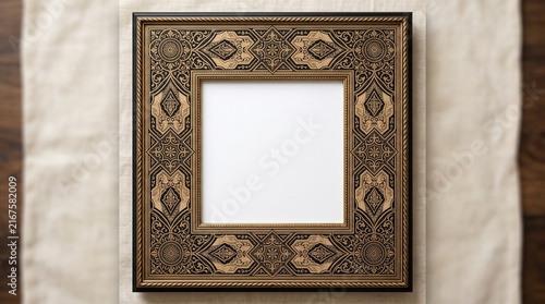 Square ornate frame with intricate geometric carvings, diamond and circular motifs, antique metallic finish, empty center on textured beige surface.