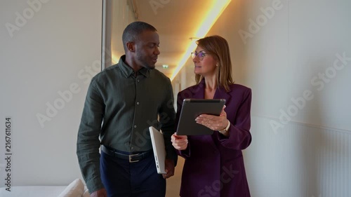 Business woman and man walking together, discussing project, carrying tablet, having conversation in office corridor. Manager and Consultant colleagues moving between meetings, busy workplace projects