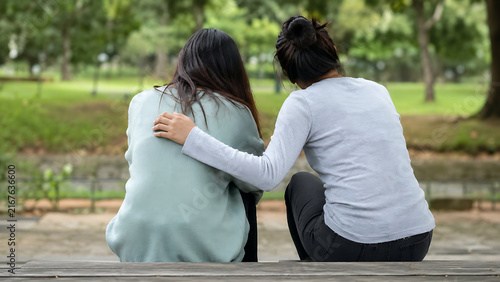 Two women sitting together in a park, one placing an arm around the other for comfort, illustrating friendship, emotional support, empathy, care, and human connection.